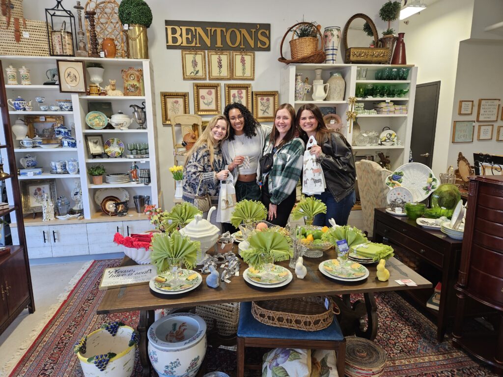 Four women stand smiling in an antique store, posing behind a table set with dinnerware and floral decorations.