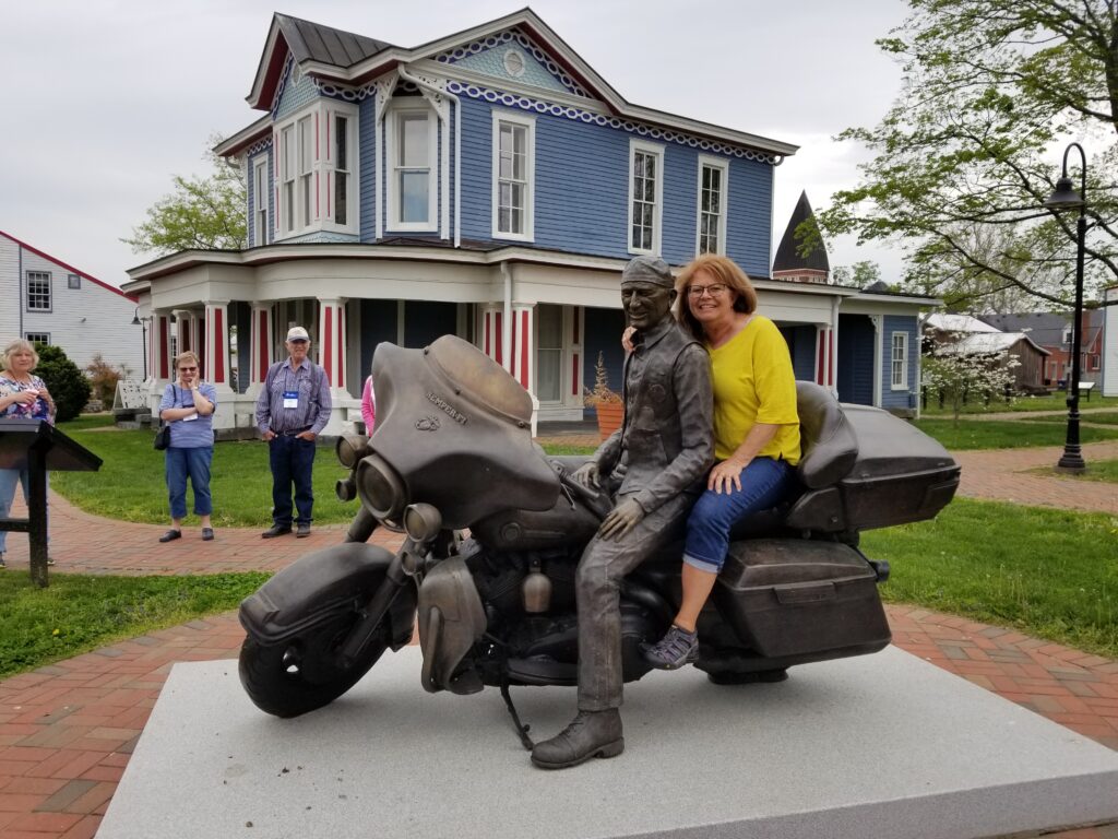 A woman sits behind a bronze statue of a man on a motorcycle in front of a blue Victorian house, while two people stand nearby.