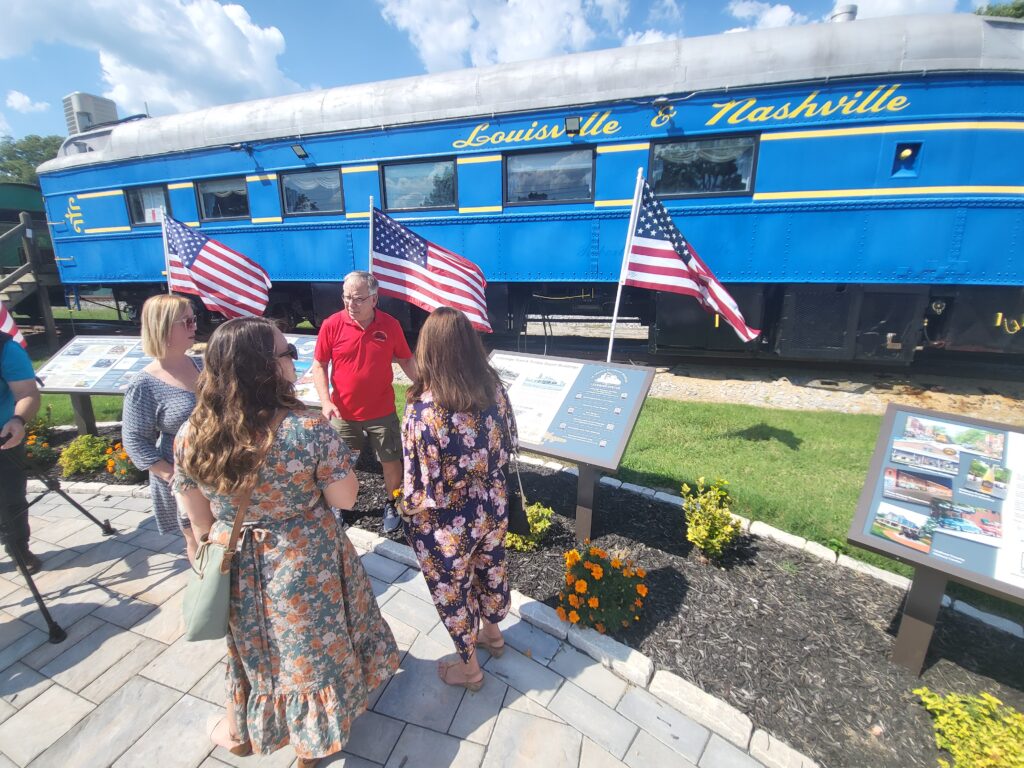 A group of people look at an information sign in front of a blue Louisville & Nashville train car flying American flags.