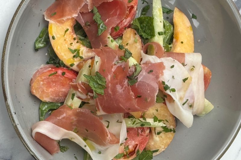 Salad of sliced peaches, prosciutto, and greens in a grey bowl.
