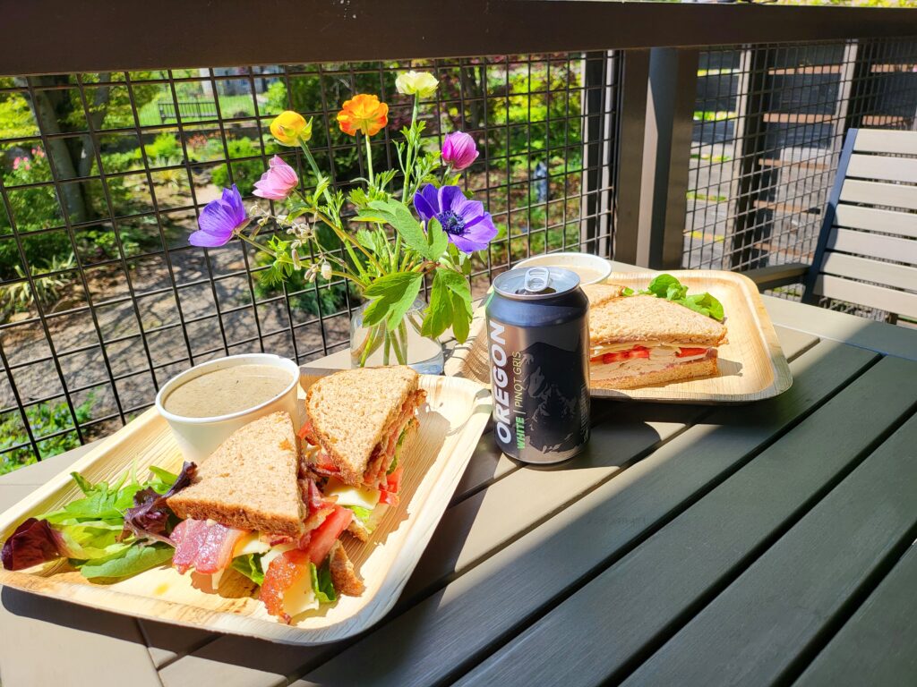 Two plates with sandwiches and small cups of soup on a patio table with a can of Oregon white pinot gris and a small vase of colorful flowers.