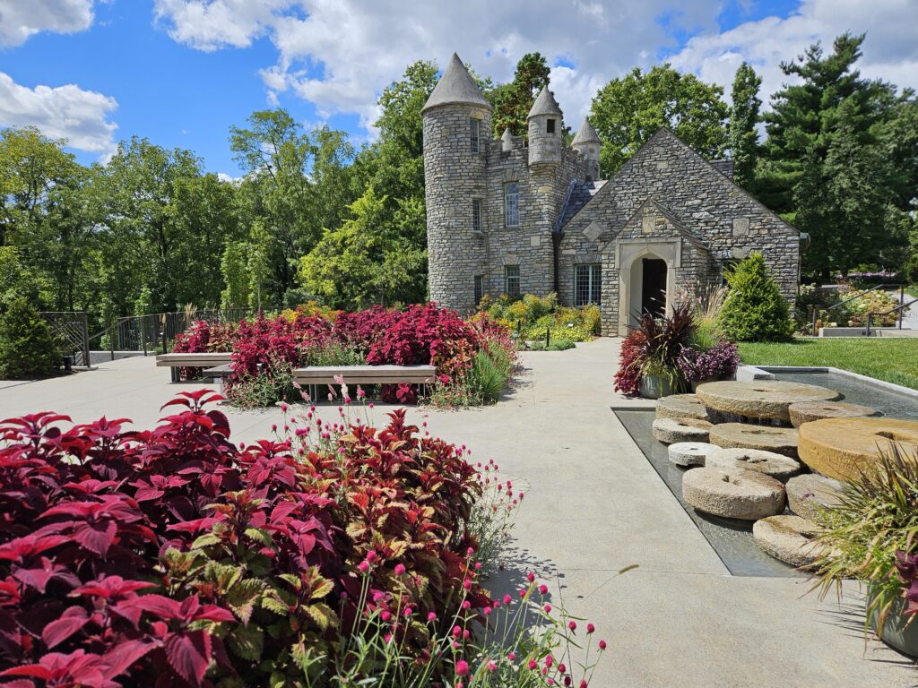 Stone castle building with turrets, surrounded by a garden with red coleus plants and a small tiered fountain.