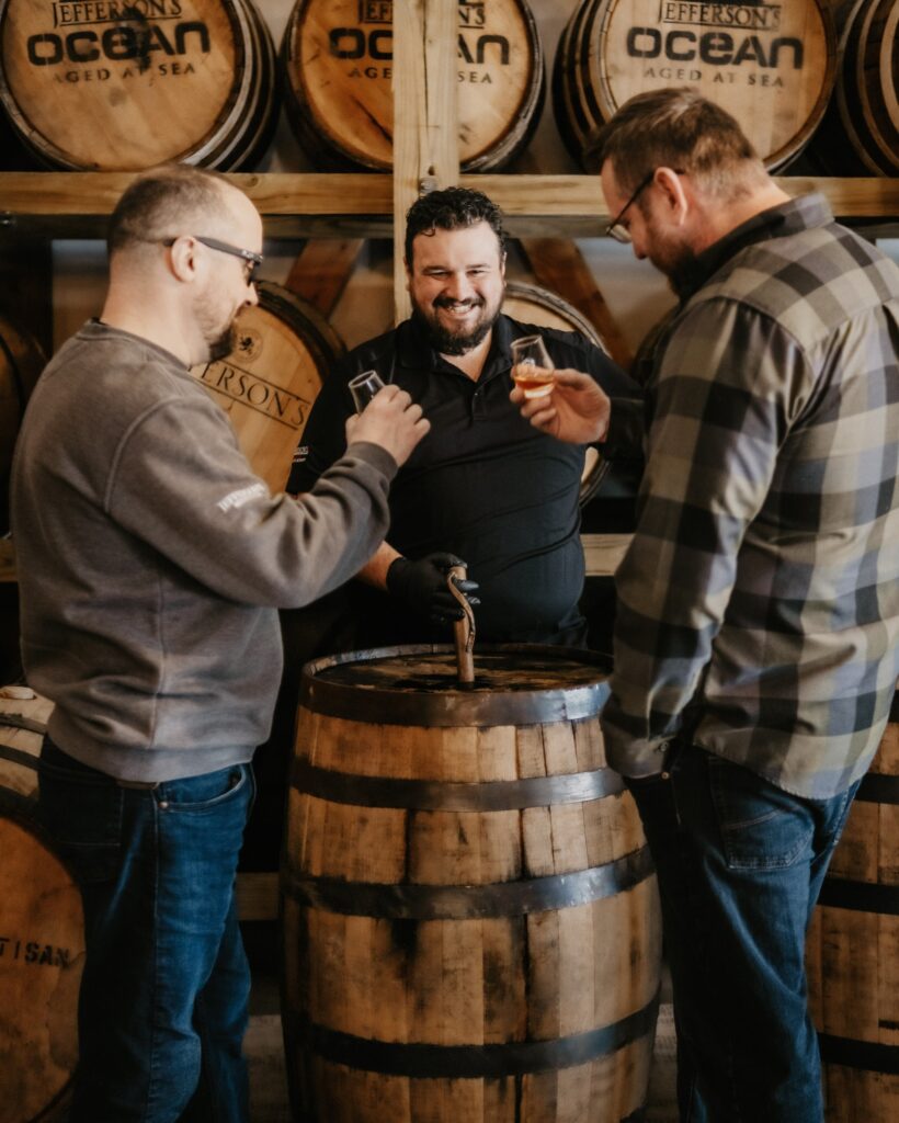 Three men tasting whiskey from small glasses in front of wooden barrels marked Jefferson’s Ocean.