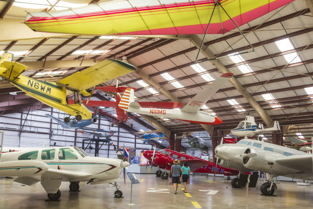 Several vintage aircraft suspended from the ceiling and parked on the hangar floor, with two people walking through the center of the display.