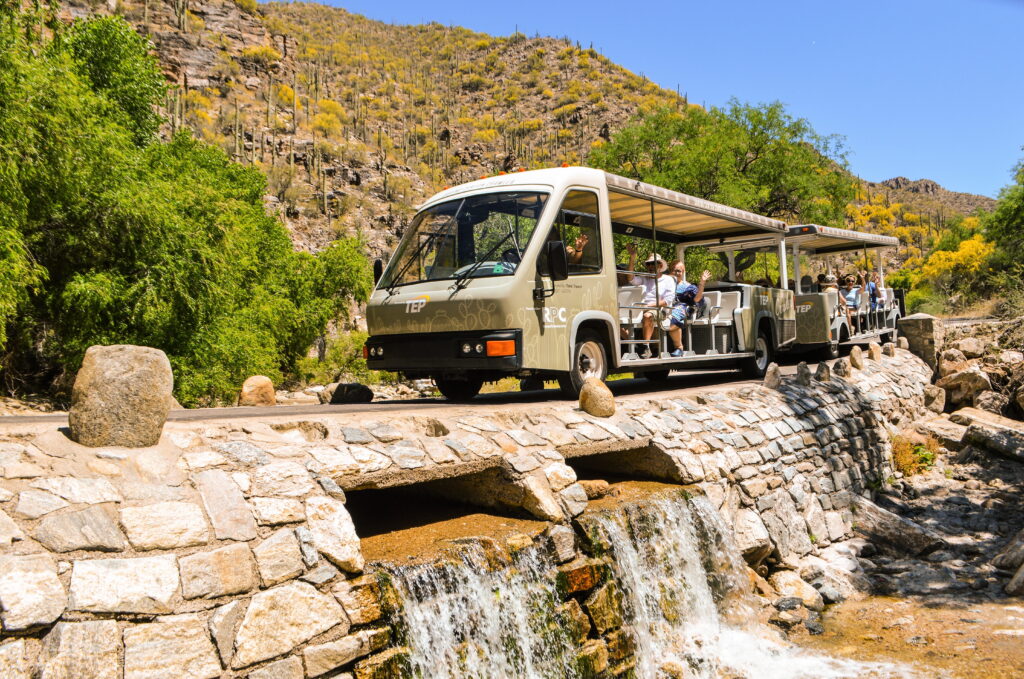 An open-air tour bus carrying passengers crosses a stone bridge over a small waterfall in a desert landscape.