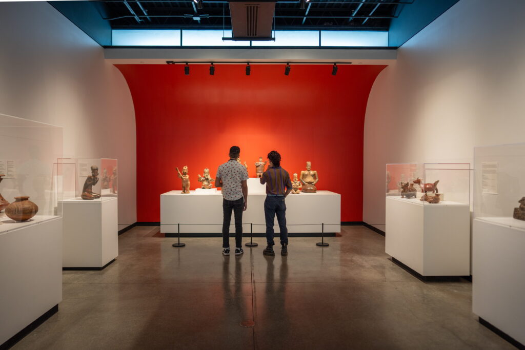 Two people stand in a museum gallery viewing a collection of ancient ceramic figurines displayed on a central pedestal against a red wall.