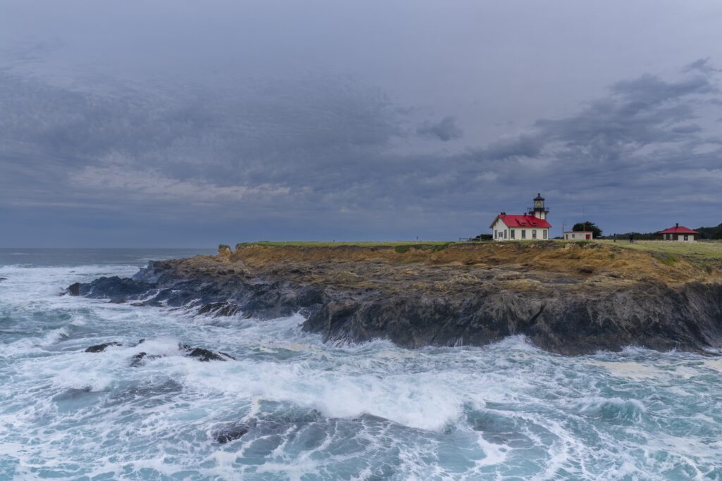 Cape Mendocino Lighthouse with a red roof perched on a rocky cliff above crashing ocean waves under a cloudy sky.