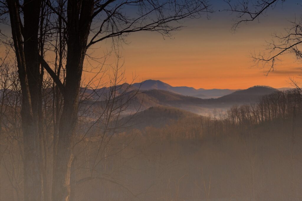 Layered mountain ridges at sunrise viewed through bare tree branches.