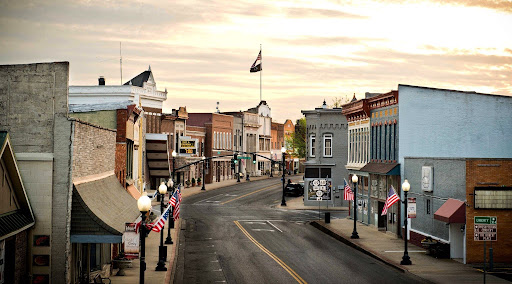 A view down a quiet small-town main street lined with multi-story brick buildings, street lamps, and American flags at sunset.