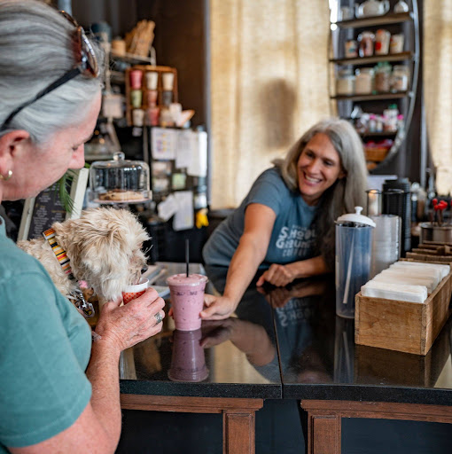 A woman feeds a small, light-colored dog from a paper cup at a coffee shop counter, while a barista smiles from across the counter.