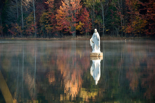 A statue of the Virgin Mary standing on a pedestal in the middle of a misty lake against a backdrop of autumn trees.